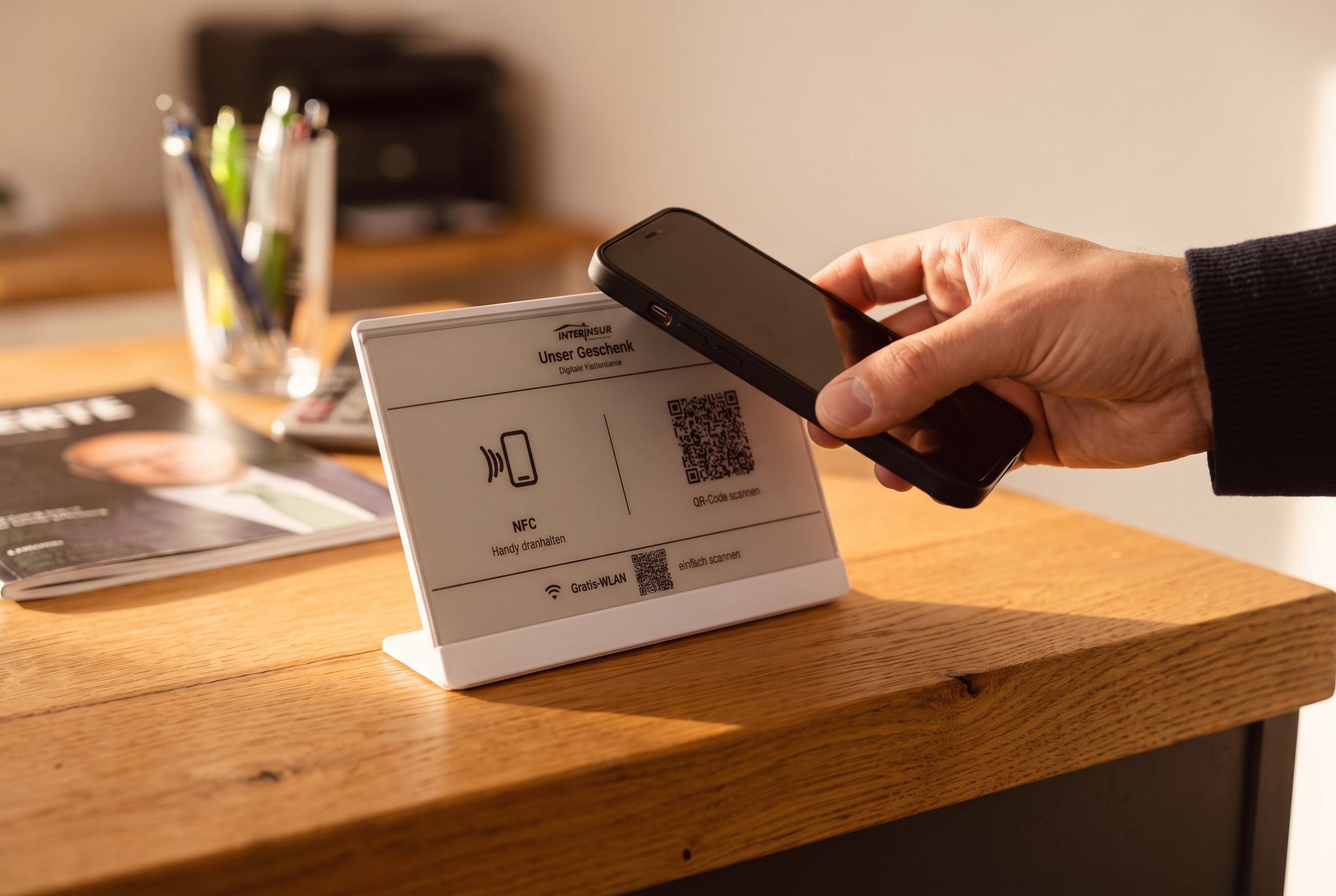 Eine Hand hält ein Smartphone an das Landingdisplay auf einem Holztresen — der Moment, in dem der Kontakt entsteht
